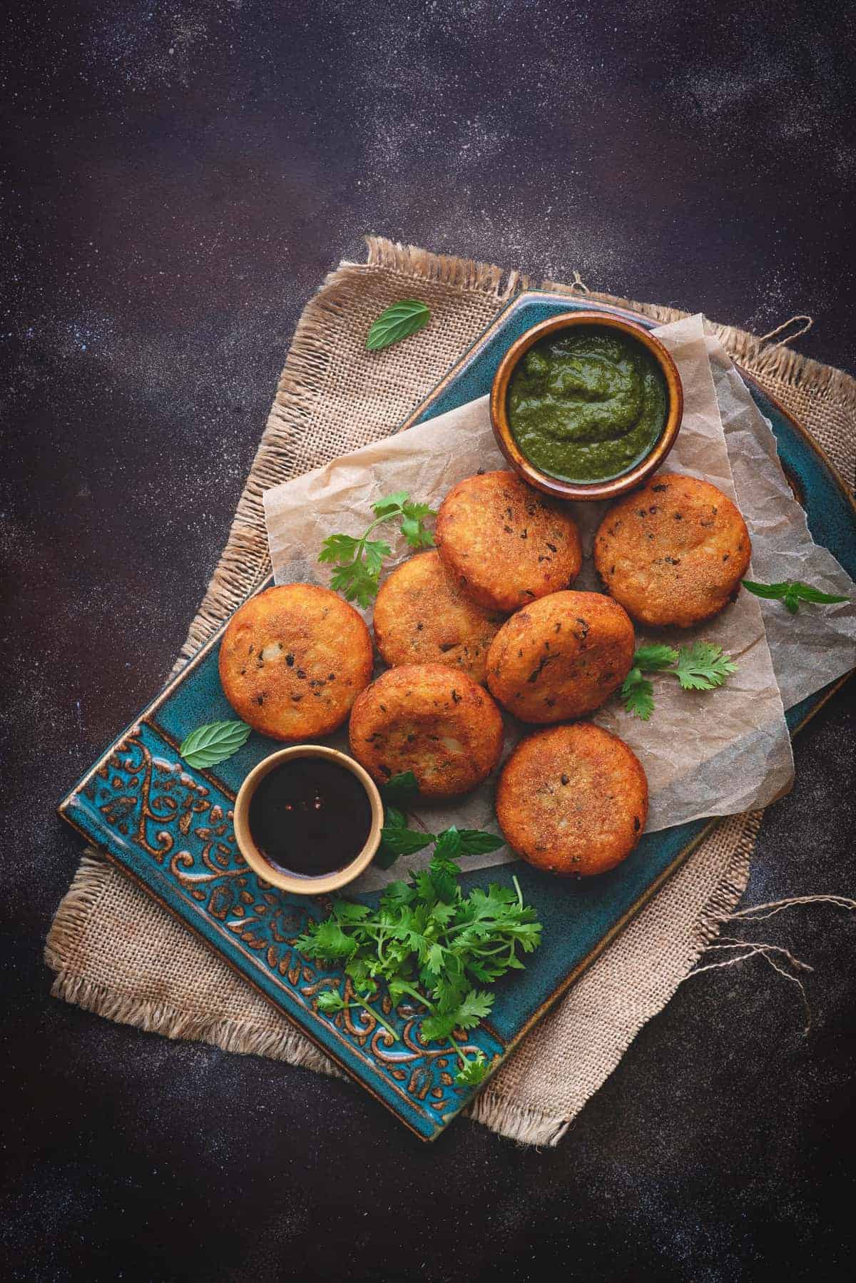 Crispy aloo patties served on blue ceramic tray, chutney in bowls on side.