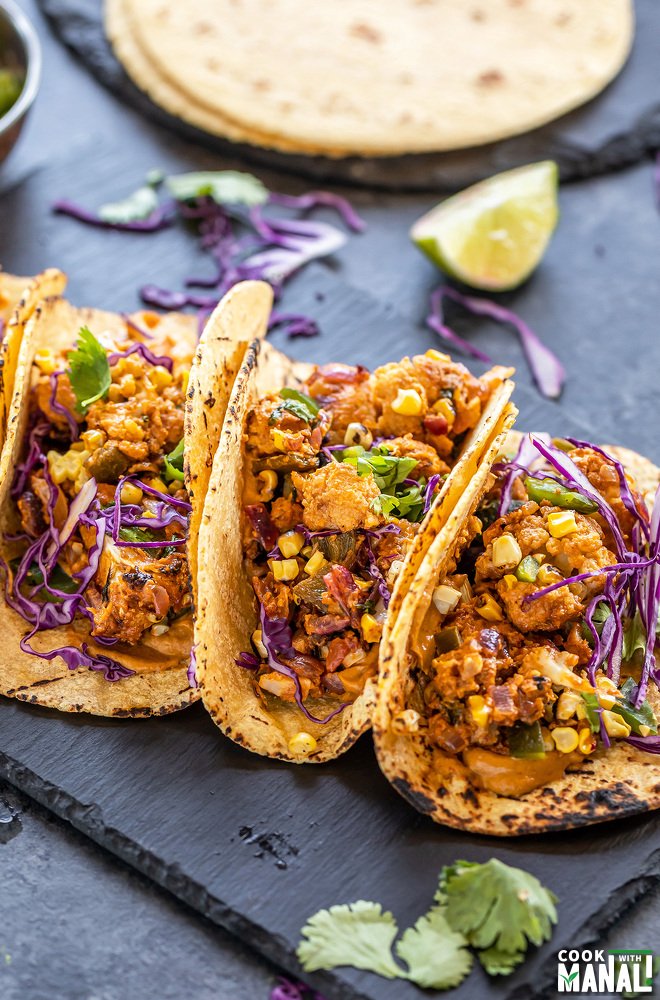 3 tacos arranged on a black board with some tortillas and lime wedges placed in the background